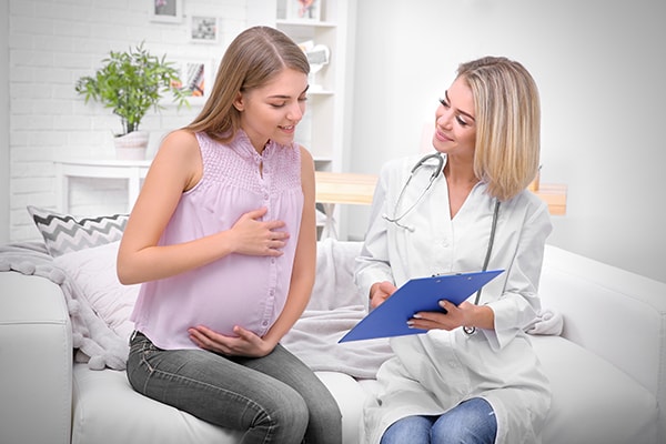 Pregnant woman wearing pastel purple sleeveless blouse holding belly talking to female doctor at home