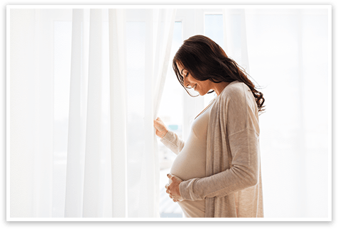 Woman holding pregnant belly wearing beige shirt and cardigan in front of white open curtain
