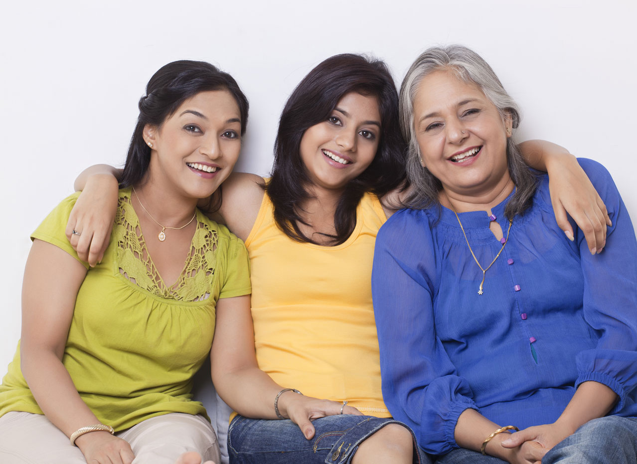 Three generations of women wearing colorful shirts holding each other in a row against light background
