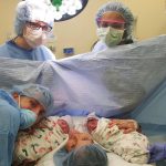 Woman and man in delivery room holding newborn twins with two others wearing blue hair-caps
