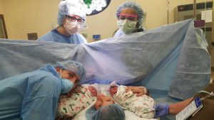 Woman and man in delivery room holding newborn twins with two others wearing blue hair-caps